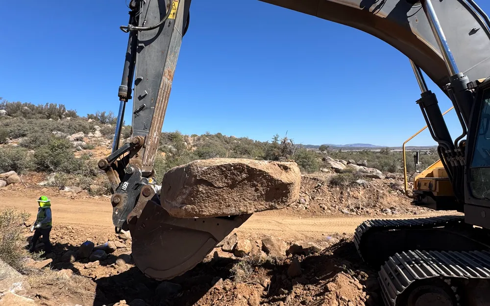 Excavator prying a large granite boulder out of rocky ground near Prescott